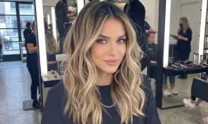 Kobieta z blond włosami i ciemnym odrostem, z niebieskimi oczami, uśmiecha się w salonie fryzjerskim.