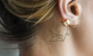 Delikatny tatuaż za uchem w kształcie żurawia origami.