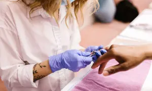 Manicurzystka w niebieskich rękawiczkach precyzyjnie usuwa skórki wokół paznokci klienta za pomocą cążków.