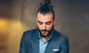 Mężczyzna z brodą i modną fryzurą samuraj, w eleganckim garniturze, spogląda w dół.