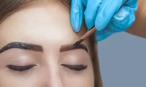 Stylist w niebieskiej rękawiczce precyzyjnie nakłada hennę na brwi, tworząc efekt brwi pudrowych.
