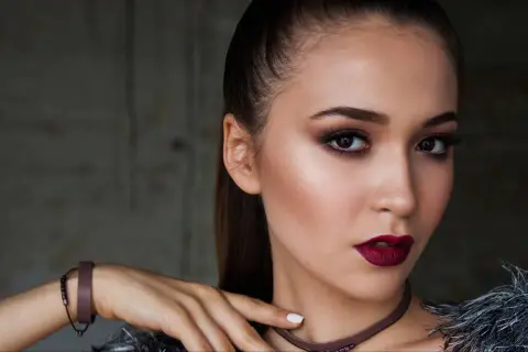 Kobieta z wyrazistym makijażem wieczorowym, ciemnymi ustami i smoky eyes. Długie włosy spięte w kucyk.