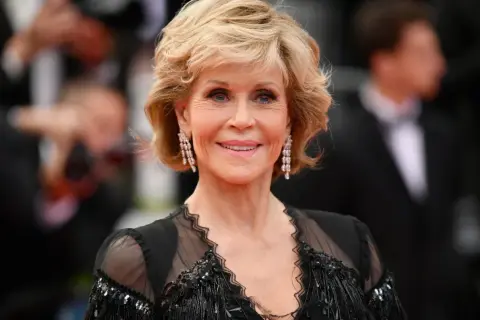 Uśmiechnięta Jane Fonda przed operacjami, w czarnej, ozdobnej sukni i długich kolczykach.