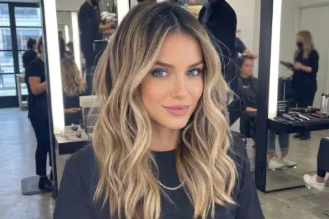 Kobieta z blond włosami i ciemnym odrostem, z niebieskimi oczami, uśmiecha się w salonie fryzjerskim.