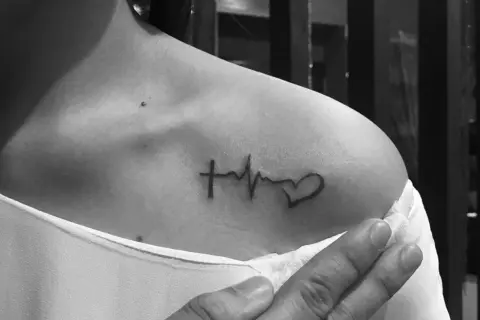 Damski tatuaż: krzyżyk, linia życia i serce na obojczyku. Symbol wiary, życia i miłości.