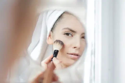 Kobieta w ręczniku na głowie nakłada puder pędzlem, patrząc w lustro. Dowiedz się, jak dobrać odcień loreal true match dla siebie.