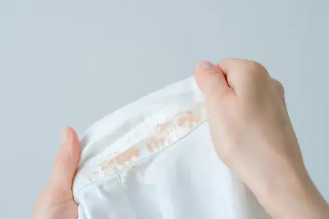 Dłonie trzymają białą koszulę z widocznymi plamami. Dowiedz się, jak usunąć plamy z podkładu, aby przywrócić ubraniu nieskazitelny wygląd.