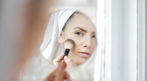 Kobieta w ręczniku na głowie nakłada puder pędzlem, patrząc w lustro. Dowiedz się, jak dobrać odcień loreal true match dla siebie.