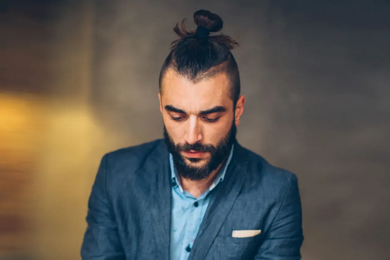 Mężczyzna z brodą i modną fryzurą samuraj, w eleganckim garniturze, spogląda w dół.
