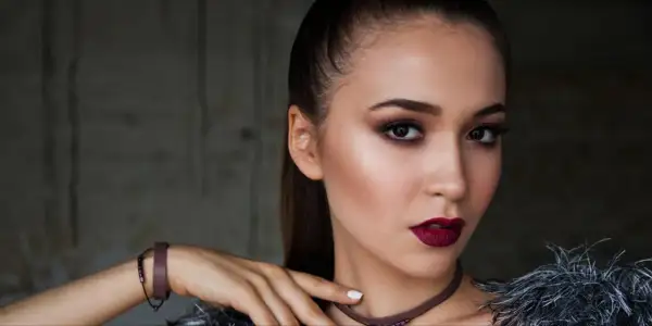 Kobieta z wyrazistym makijażem wieczorowym, ciemnymi ustami i smoky eyes. Długie włosy spięte w kucyk.