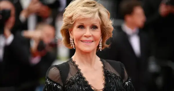 Uśmiechnięta Jane Fonda przed operacjami, w czarnej, ozdobnej sukni i długich kolczykach.