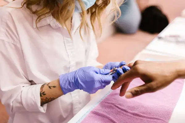 Manicurzystka w niebieskich rękawiczkach precyzyjnie usuwa skórki wokół paznokci klienta za pomocą cążków.