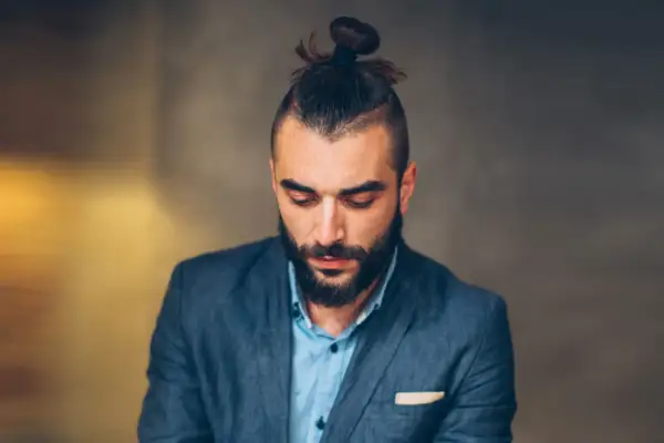 Mężczyzna z brodą i modną fryzurą samuraj, w eleganckim garniturze, spogląda w dół.