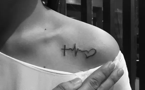 Damski tatuaż: krzyżyk, linia życia i serce na obojczyku. Symbol wiary, życia i miłości.