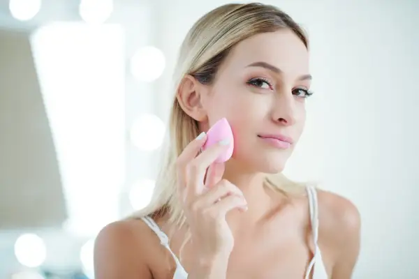 Blondynka z naturalnym pięknem, używa różowej gąbeczki do makijażu. Efekt make up no make up.