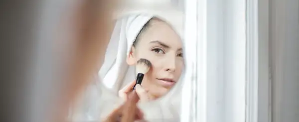 Kobieta w ręczniku na głowie nakłada puder pędzlem, patrząc w lustro. Dowiedz się, jak dobrać odcień loreal true match dla siebie.